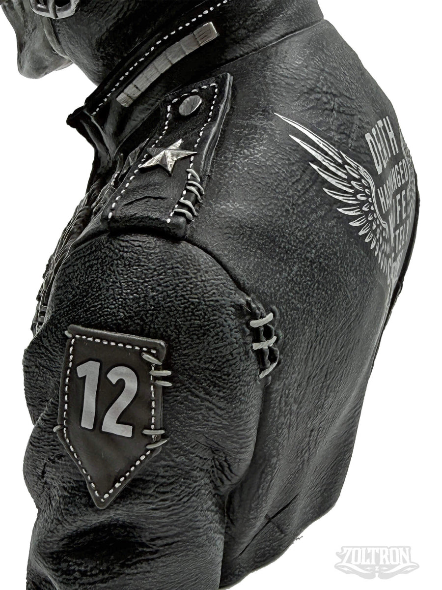 A side view close-up of a highly detailed resin and soapstone sculpture by artist Zoltron, featuring a highly detailed skull figurine wearing a cracked leather aviator helmet, a weathered black leather jacket, war medallions and adornments, smoking a cigarette, set against a plain white background.