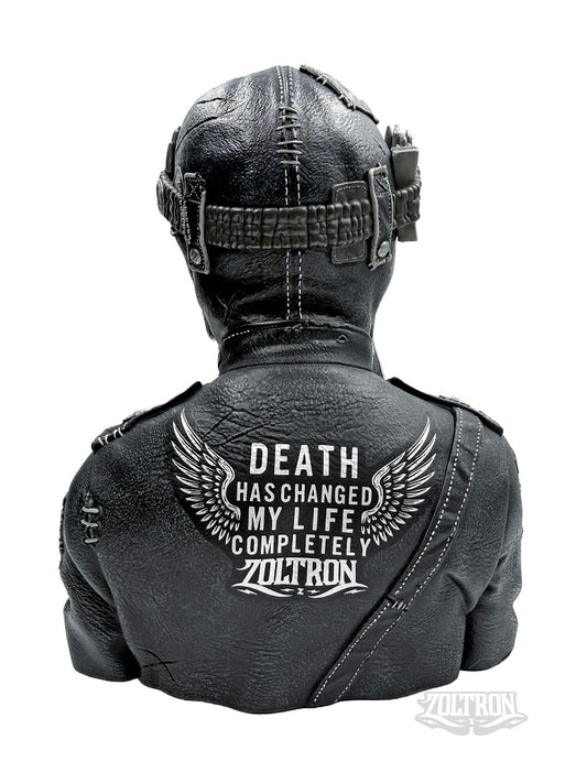 A backview close-up of a highly detailed resin and soapstone sculpture by artist Zoltron, featuring a highly detailed skull figurine wearing a cracked leather aviator helmet, a weathered black leather jacket adorned with the phrase DEATH HAS CHANGED MY LIFE COMPLETELY. war medallions and adornments, stitching and distress from time,  set against a plain white background.